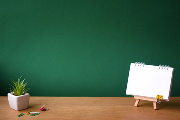 Back to school concept, open notebook on miniature easel and small succulent in a white pot on wooden surface on the backdrop of a clean green chalk board, selective focus, copy space