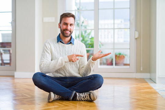 Handsome man wearing casual sweater sitting on the floor at home amazed and smiling to the camera while presenting with hand and pointing with finger.