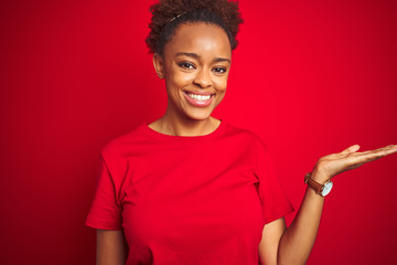 Young beautiful african american woman with afro hair over isolated red background smiling cheerful presenting and pointing with palm of hand looking at the camera.