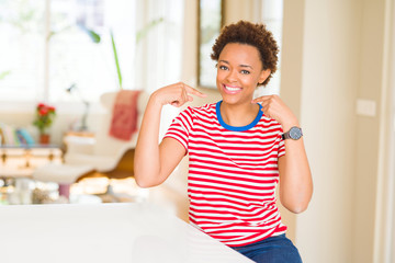 Young beautiful african american woman at home looking confident with smile on face, pointing...