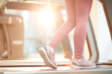 Sports background of unrecognizable woman running on treadmill in gym lit by sunlight, copy space