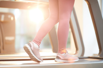Sports background of slender female legs running on treadmill in gym lit by sunlight, copy space