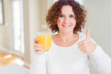 Senior woman driking a glass of fresh orange juice happy with big smile doing ok sign, thumb up with fingers, excellent sign