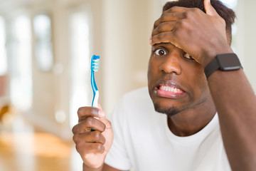 African american man holding toothbrush stressed with hand on head, shocked with shame and surprise...