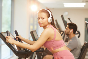 Waist up portrait of young woman wearing headphones looking at camera while exercising in gym, copy space