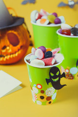 Halloween trick or treat sweets. Paper cups with colourful candies inside and paper silhouettes of bats, ghosts, witch on bright yellow background. Spooky holiday symbols. Vertical card. Copy space.