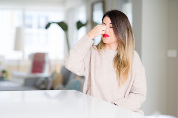 Young beautiful woman wearing winter sweater at home smelling something stinky and disgusting,...