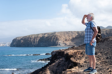 An attractive senior man casual dressed standing in front to the sea and looking away. White hair and beard. Smiling. Sunrise. Blue ocean and sky on background