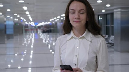 Positive business woman look straight to camera with smile, blurred airport passage on background. Friendly lady hold smartphone in hands, half-length portrait shot