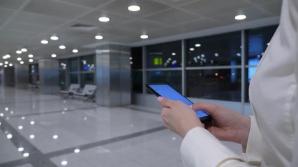 Woman texting on phone on the go, closeup shot of mobile device in hands. Passenger lady walk at empty airport passage in late evening.