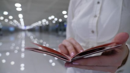 Closeup shot of passport, woman flip pages and look to visa pages, stamps and marks, blurred empty airport hall seen on background. Passenger lady wear white shirt