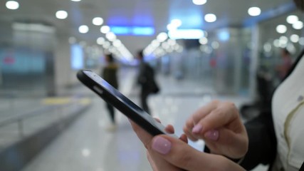 Smartphone in woman hands, she type message using finger and pressing letter buttons at on-screen keyboard. Blurred airport passage on background, people silhouettes