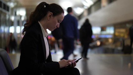 Woman look through images on smartphone, sit at airport hall, blurred background. Passenger lady spend time waiting for boarding at international terminal