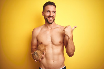 Young handsome shirtless man holding exotic tropical coconut over isolated yellow background pointing and showing with thumb up to the side with happy face smiling