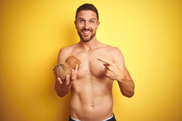 Young handsome shirtless man holding exotic tropical coconut over isolated yellow background very happy pointing with hand and finger