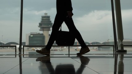 Woman with handbag walk against terminal window, black silhouette of passenger legs, slow motion shot. Air traffic control tower seen outside, dim illumination at green concourse