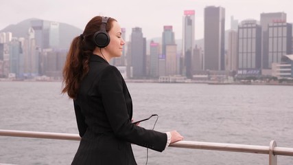 Business woman stay with eyes closed, listen music from smartphone at Victoria Harbour embankment, against beautiful Hong Kong island waterfront. Professional lady enjoy vacant time during duty trip