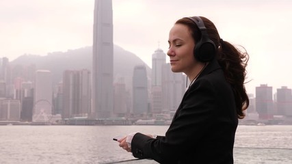Woman in black suit stay with eyes closed on embankment, listen music, half length shot. Long hair fly on breeze, business lady enjoy and rest in short pause after work
