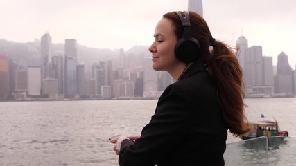 Woman in black suit rest at city embankment, listen music, stay with eyes closed against Victoria Harbour. Hong Kong island buildings seen on background