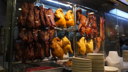 Roasted and boiled whole chicken and pork pieces, pre-cooked meat hang at Chinese restaurant stall. Unidentified cook man prepare some meals, seen behind
