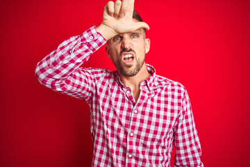 Young handsome man over red isolated background making fun of people with fingers on forehead doing...