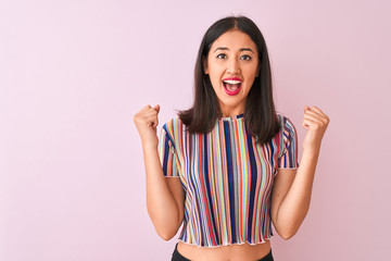 Young chinese woman wearing colorful striped t-shirt standing over isolated pink background celebrating surprised and amazed for success with arms raised and open eyes. Winner concept.