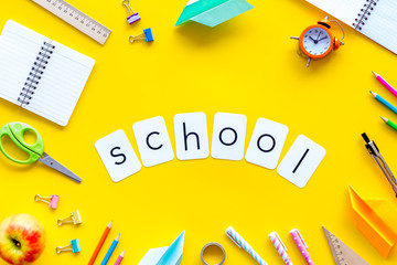Education concept with school word and frame of stationery with notebook, pens, clock on yellow background top view
