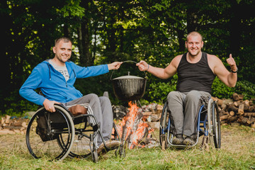 Two disabled men resting in a campsite with friends. Wheelchair in the forest on the background of...