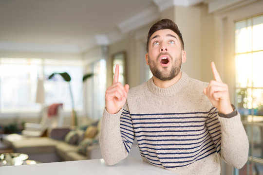 Young handsome man at home amazed and surprised looking up and pointing with fingers and raised arms.