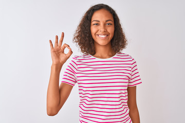 Young brazilian woman wearing pink striped t-shirt standing over isolated white background smiling positive doing ok sign with hand and fingers. Successful expression.