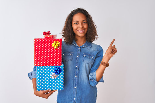 Young brazilian woman holding birthday gifts standing over isolated white background very happy pointing with hand and finger to the side