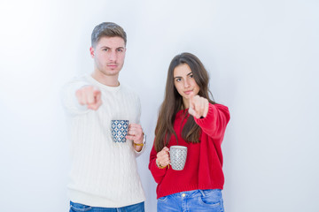 Beautiful young couple over white isolated background drinking a cup of coffee pointing with finger to the camera and to you, hand sign, positive and confident gesture from the front