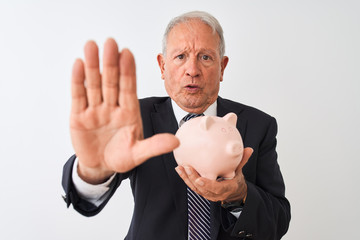 Senior grey-haired businessman holding piggy bank standing over isolated white background with open hand doing stop sign with serious and confident expression, defense gesture