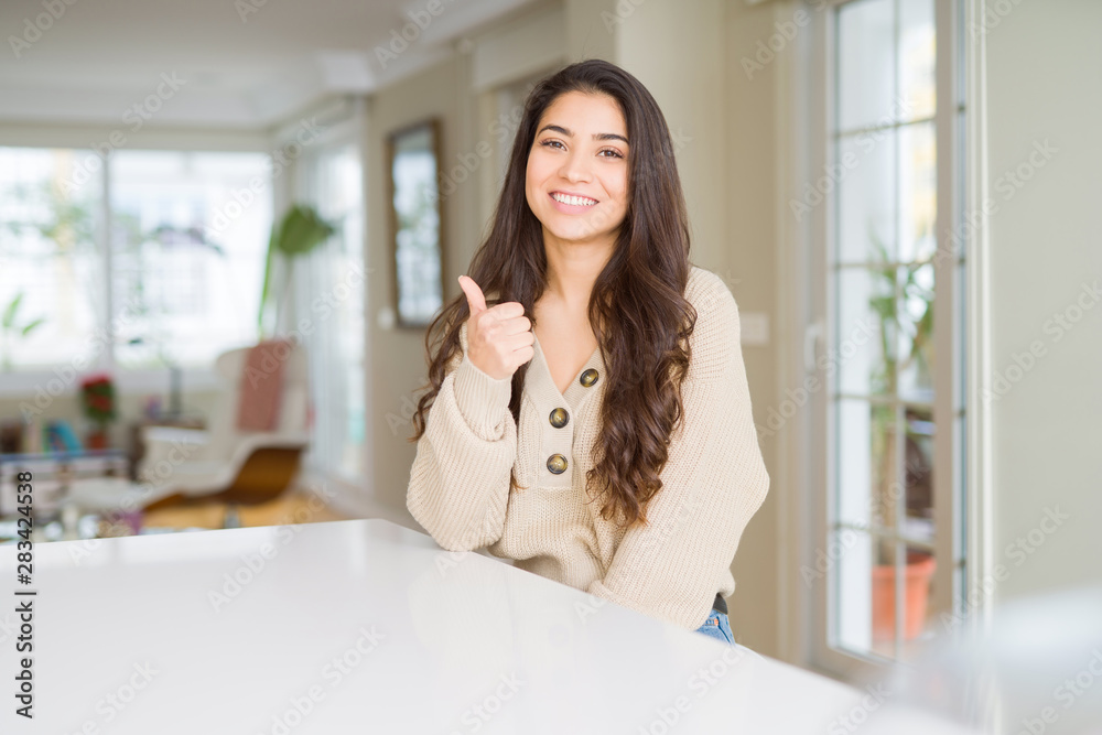 Wall mural young beautiful woman at home doing happy thumbs up gesture with hand. approving expression looking 