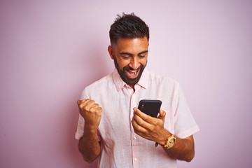 Young indian man using smartphone standing over isolated pink background screaming proud and...