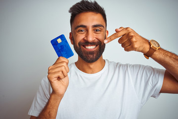 Young indian customer man holding credit card standing over isolated white background very happy...