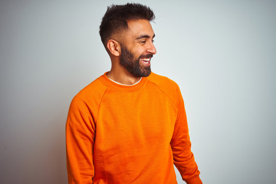 Young indian man wearing orange sweater over isolated white background looking away to side with smile on face, natural expression. Laughing confident.