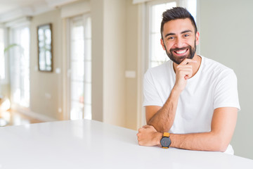 Handsome hispanic man casual white t-shirt at home looking confident at the camera with smile with crossed arms and hand raised on chin. Thinking positive.