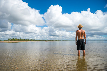 Tourist wearing a straw sun hat standing in calm shallow waters on the shore of a sunny palm-lined tropical island beach 