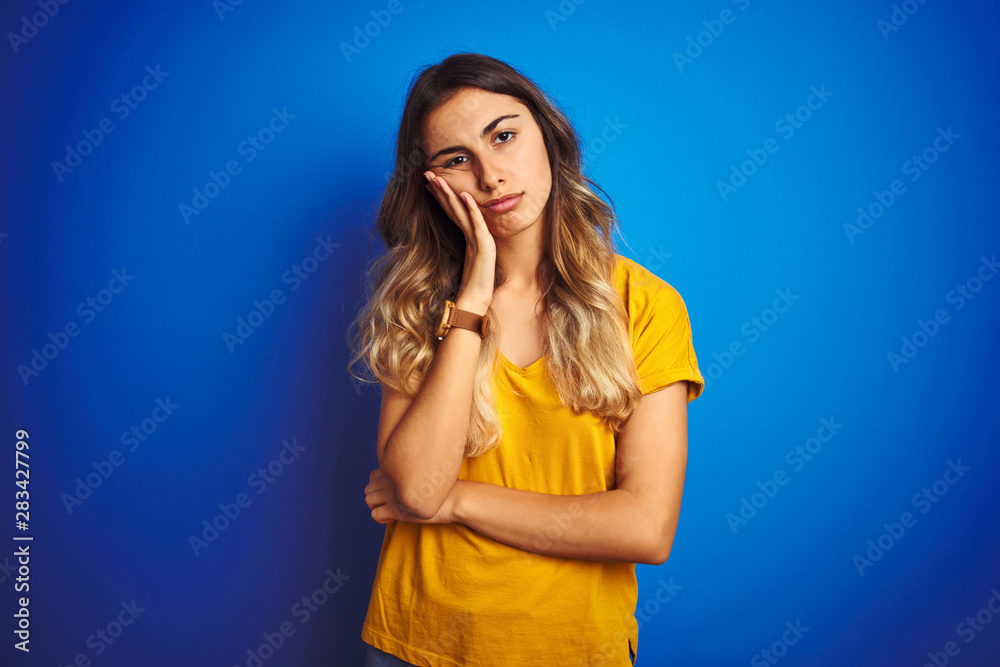 Sticker Young beautiful woman wearing yellow t-shirt over blue isolated background thinking looking tired and bored with depression problems with crossed arms.