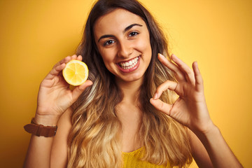Young beautiful woman holding half lemon over yellow isolated background doing ok sign with fingers, excellent symbol