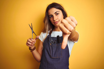 Young beautiful hairdresser woman holding scissors over yellow isolated background with angry face,...