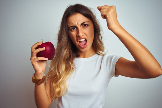 Young beautiful woman eating red apple over grey isolated background annoyed and frustrated shouting with anger, crazy and yelling with raised hand, anger concept