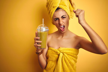 Young beautiful woman wearing a towel drinking detox juice over yellow isolated background annoyed and frustrated shouting with anger, crazy and yelling with raised hand, anger concept