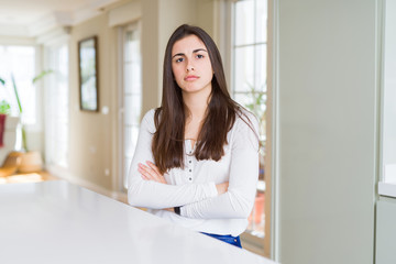 Beautiful young woman sitting on white table at home skeptic and nervous, disapproving expression on face with crossed arms. Negative person.