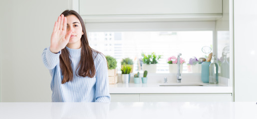 Wide angle picture of beautiful young woman sitting on white table at home doing stop sing with palm of the hand. Warning expression with negative and serious gesture on the face.