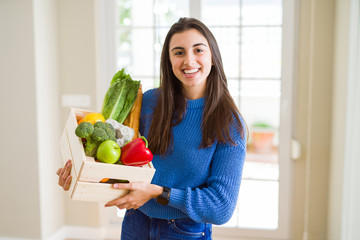 Beautiful young woman holding wooden box full of healthy groceries with a happy face standing and smiling with a confident smile showing teeth