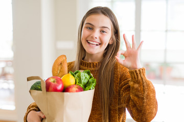 Beautiful young girl holding paper bag of fresh groceries doing ok sign with fingers, excellent symbol