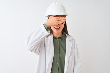 Young chinese engineer woman wearing coat helmet glasses over isolated white background smiling and laughing with hand on face covering eyes for surprise. Blind concept.