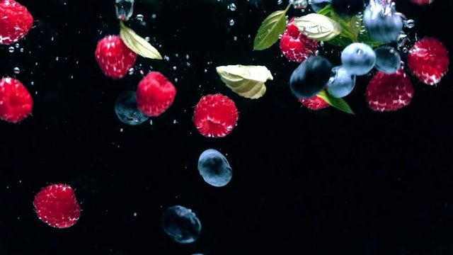 Fresh raspberries with blueberries and fresh mint leaves fall on a black background into the water, splashing on a black background. Drop fresh fruits and berries into the water. Healthy eating, slow